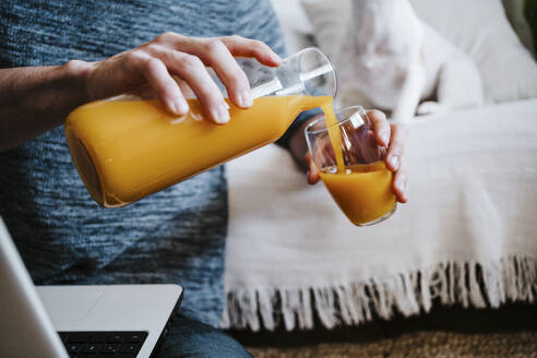 Mann gießt Saft in ein Glas, während er zu Hause am Laptop sitzt - EBBF01776