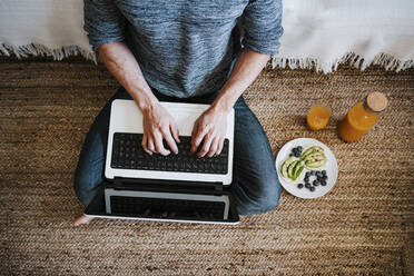 Man working on laptop while sitting by plate of fruit and juice at home - EBBF01779