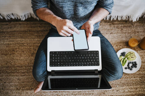 Mann mit Laptop, der ein Mobiltelefon benutzt, während er beim Frühstück zu Hause auf dem Boden sitzt - EBBF01781