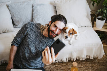 Lächelnder Mann nimmt Selfie mit Hund durch Handy, während er zu Hause sitzt - EBBF01783