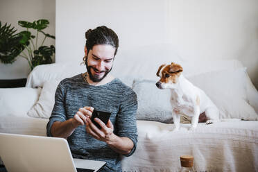 Mann mit Laptop, der ein Mobiltelefon benutzt, während er bei seinem Hund zu Hause sitzt - EBBF01784