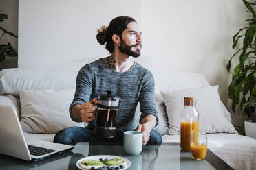 Mann mit Kaffeemaschine, der wegschaut, während er beim Frühstück auf dem Tisch zu Hause sitzt - EBBF01786
