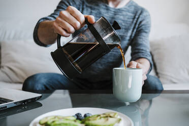 Mann hält Kaffeemaschine und gießt Kaffee in Kaffeetasse zu Hause sitzend - EBBF01788