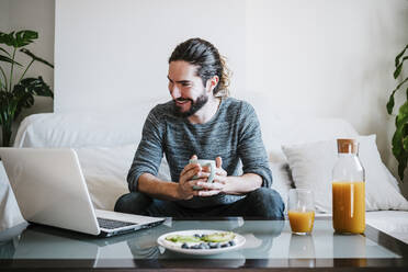 Junger Mann mit Kaffeetasse und Laptop, während er zu Hause auf dem Sofa sitzt und frühstückt - EBBF01791