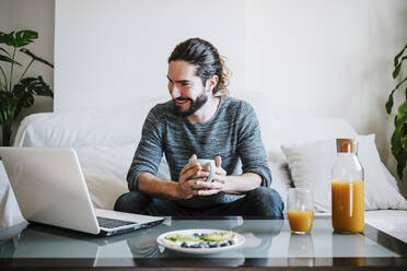 Young man with coffee cup using laptop while having breakfast sitting on sofa at home - EBBF01791