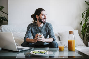 Smiling man with coffee cup looking away while having breakfast sitting on sofa at home - EBBF01792