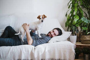 Glücklicher Mann lächelt, während er mit seinem Haustier spielt, das auf dem Sofa zu Hause liegt - EBBF01796