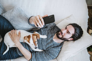Junger Mann, der ein Selfie mit seinem Handy macht, während er mit seinem Hund zu Hause auf dem Sofa liegt - EBBF01798