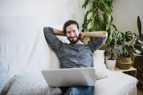 Lächelnder Mann mit Laptop, der zu Hause auf dem Sofa sitzt - EBBF01807