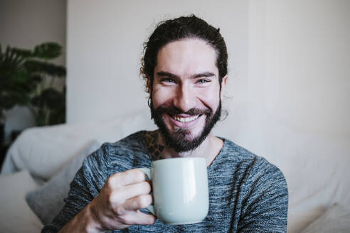 Junger Mann lächelnd beim Kaffeetrinken zu Hause sitzend - EBBF01809