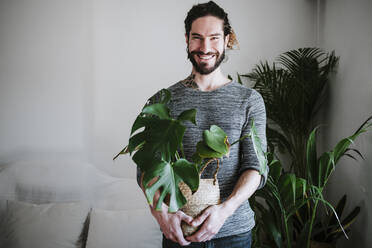 Junger Mann lächelt, während er eine Monstera-Pflanze zu Hause hält - EBBF01819