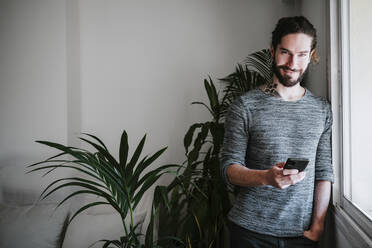 Young man smiling while using mobile phone leaning by window at home - EBBF01820