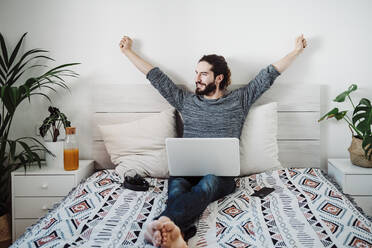 Mann beim Dehnen mit Laptop auf dem Bett sitzend zu Hause - EBBF01830