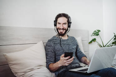 Lächelnder Mann mit Kopfhörern, der ein Mobiltelefon benutzt, während er mit einem Laptop auf dem Bett zu Hause sitzt - EBBF01831