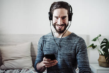 Lächelnder Mann mit Kopfhörern, der ein Mobiltelefon benutzt, während er zu Hause auf dem Bett sitzt - EBBF01834
