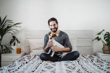 Hübscher Mann sitzt mit Hund auf dem Bett zu Hause - EBBF01836