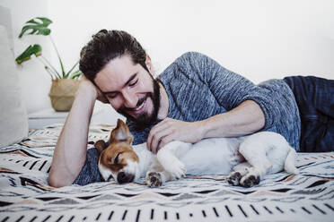 Young man smiling while looking at dog sleeping on bed at home - EBBF01839