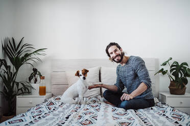 Gut aussehender Mann spielt mit Hund, während er zu Hause auf dem Bett sitzt - EBBF01843