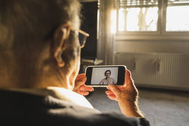 Ein älterer Mann lässt sich von einer Allgemeinmedizinerin per Videoanruf über ein Smartphone im Wohnzimmer beraten - UUF22274