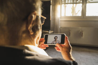 Senior man taking advice from female general practitioner on video call through smart phone in living room - UUF22274