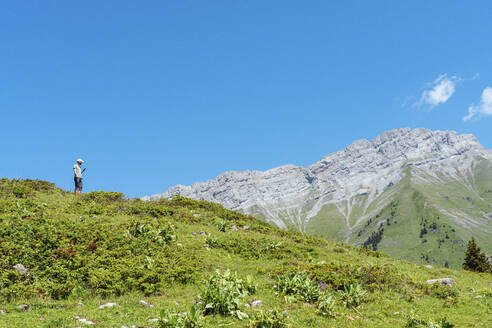 Mittlerer erwachsener Mann, der ein Mobiltelefon benutzt, während er auf einer Wiese am Col des Aravis steht, Haute-Savoie, gegen den Himmel, Frankreich - GEMF04482