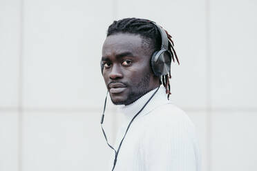 Young man listening music through headphones against white wall - EBBF01845