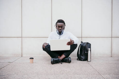 Junger Mann arbeitet am Laptop, während er auf dem Gehweg vor einer weißen Wand sitzt - EBBF01867