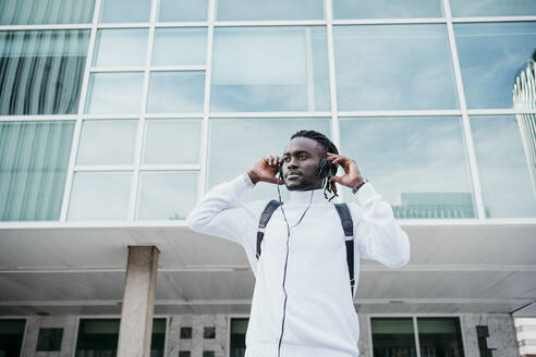 Junger Geschäftsmann hört Musik über Kopfhörer an einem Gebäude in der Stadt - EBBF01877