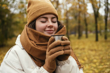 Smiling young woman with eyes closed smelling tea in autumn park - OYF00276