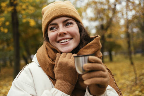 Glückliche junge Frau schaut weg, während sie eine isolierte Tasse im Herbstpark hält - OYF00277