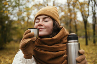 Junge Frau mit geschlossenen Augen riecht Tee im herbstlichen Park - OYF00279