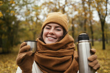 Smiling young woman with insulated drink container at autumn park - OYF00280