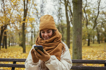Smiling young woman using mobile phone while sitting on bench at autumn park - OYF00281