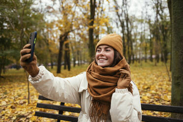 Happy young woman taking selfie while sitting on bench at autumn park - OYF00283