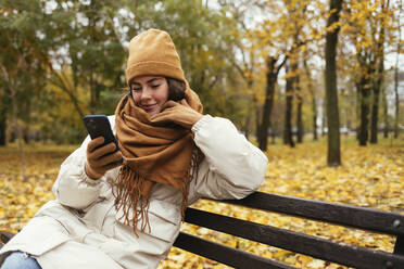 Lächelnde junge Frau mit Hand am Kinn, die im herbstlichen Park ein Mobiltelefon benutzt - OYF00284
