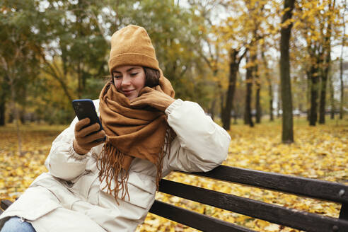 Lächelnde junge Frau mit Hand am Kinn, die im herbstlichen Park ein Mobiltelefon benutzt - OYF00284