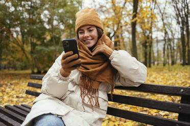 Happy young woman with scarf around neck using smart phone on bench at autumn park - OYF00285