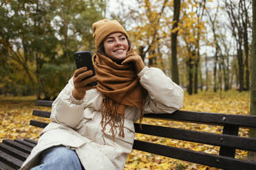 Smiling woman day dreaming while holding mobile phone in autumn park - OYF00286