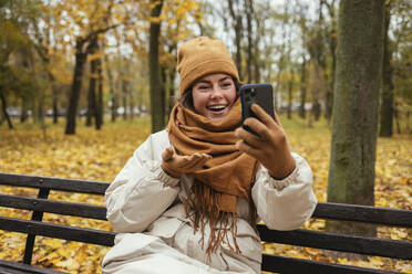Fröhliche junge Frau bei einem Videogespräch, während sie auf einer Bank im Herbstpark sitzt - OYF00287