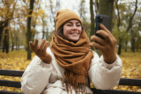 Lächelnde junge Frau, die mit der Hand gestikuliert, während sie einen Videoanruf im Herbstpark führt - OYF00289