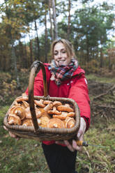 Smiling woman showing basket full of mushrooms in forest in autumn - JAQF00018