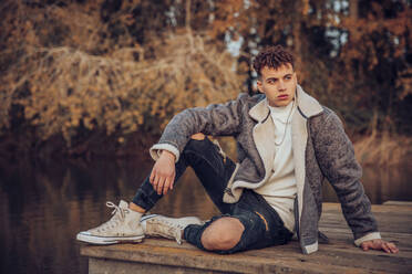 Young man wearing jacket sitting on jetty against lake at autumn forest - ACPF00971