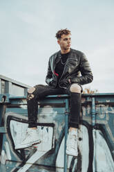 Young man with hands in pockets sitting on metal container full of garffiti against sky - ACPF00973