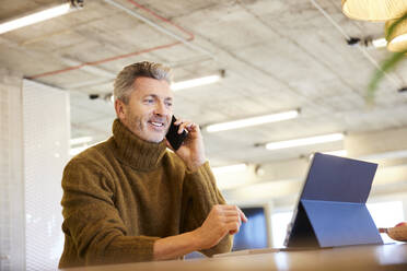 Man talking on mobile phone while using digital tablet sitting at home - FMKF06836