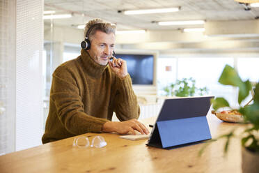 Businessman wearing headset using digital tablet while sitting at office - FMKF06848