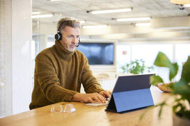Businessman wearing headset talking while working on digital tablet at office - FMKF06849