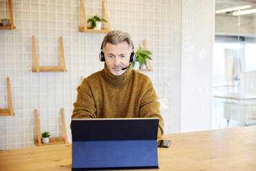 Male businessman wearing headset working on digital tablet while sitting at office - FMKF06850