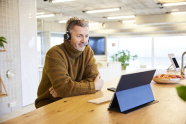 Mature businessman wearing headset working on digital tablet while sitting at office - FMKF06854