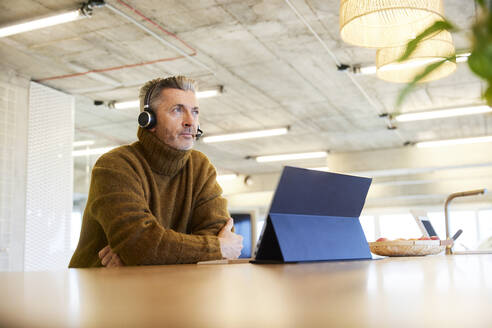 Thoughtful businessman with headset and digital tablet looking away while sitting at office - FMKF06855