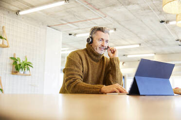Male businessman wearing headset talking while working on digital tablet at office - FMKF06856